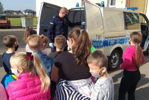 Policjant prewencji z Działdowa na placu przed budynkiem szkoły prezentuje dzieciom ze Szkoły Podstawowej w Księżym Dworze policyjny radiowóz