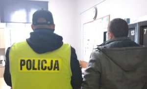 zatrzymany mężczyzna podejrzany o zabójstwo 30-latki znajdujący się obok prowadzącego do policjanta, który ma na sobie kamizelkę z napisem Policja