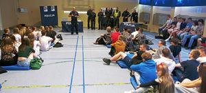 Zastępca Komendanta Powiatowego Policji w Działdowie podczas pogadanki z uczniami, w tle funkcjonariusze i banery policyjne